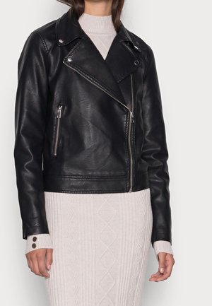 Kvinde iført en sort læderbikerjakke over en lysebeige strikket kjole med knappede ærmekanter, stående foran en ensfarvet baggrund.