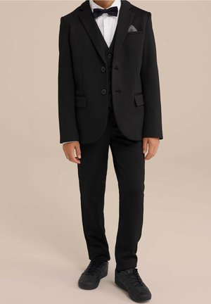 Traje negro de tres piezas con acabado texturizado, camisa blanca, pajarita negra y un pañuelo de bolsillo. Combinado con zapatillas negras.