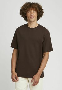 T-shirt marron texturé à manches courtes et col rond, avec un motif côtelé. Modèle portant un pantalon clair.