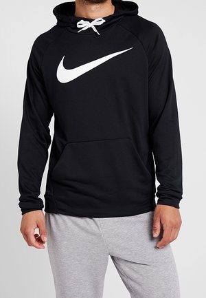 Mand iført en sort Nike hættetrøje med hvidt logo og hvide snore, kombineret med lysegrå joggingbukser mod en ensfarvet baggrund.