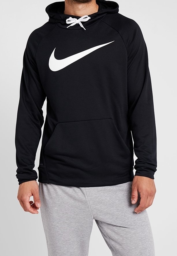 Homme portant un sweat à capuche noir Nike avec logo blanc et cordon blanc, associé à un pantalon de survêtement gris clair sur un fond uni.