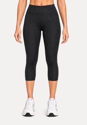 Femme portant des leggings courts noirs à imprimé léopard et des chaussures de sport blanches, debout avec les bras relâchés le long du corps.
