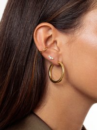 Oreille droite avec trois boucles d'oreilles en or : un grand anneau, un clou à trois pierres et un bracelet torsadé, encadrée par des cheveux bruns raides.