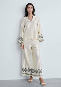 Silk cream top with V-neck and long sleeves, features black geometric embroidery. Paired with matching wide-leg pants, also silk.