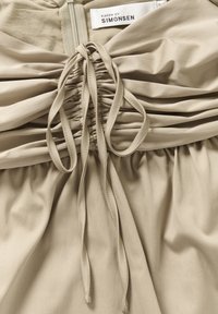 Tessuto beige con design arricciato, caratterizzato da cordini ed una zip sul retro. Texture morbida con una leggera lucentezza. Etichetta del marchio visibile.