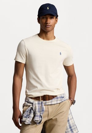 T-shirt di cotone beige con colletto rotondo, caratterizzata da un logo ricamato blu. Abbinata a pantaloni khaki e una camicia a quadri blu annodata in vita.