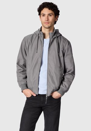 Nuori mies, jolla on kiharat hiukset, pukeutunut harmaaseen vetoketjulliseen huppariin vaaleansinisen paidan ja mustien housujen päällä, kädet taskuissa, neutraali tausta.