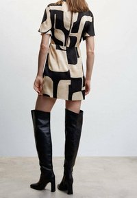 Krátké šaty v béžovém a černém geometrickém vzoru, se zadním výkopem. Spárované s elegantními černými vysokými kozačkami na podpatku.