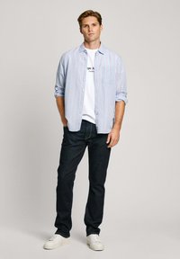 Light blue striped button-up shirt over a white t-shirt, paired with dark jeans and white sneakers. Straight-leg design with a front pocket.