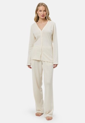 Mujer rubia de pie descalza con top de manga larga acanalado crema con botones y pantalones anchos a juego sobre fondo blanco.