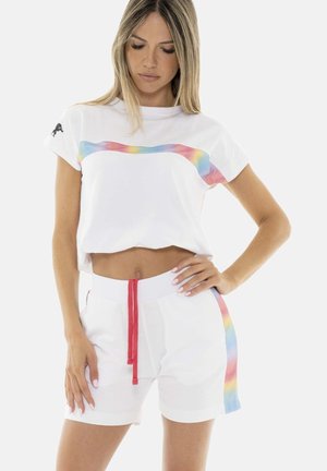 Femme portant un t-shirt court blanc et un short à rayures arc-en-ciel, posant avec une main sur la hanche et l'autre le long du corps.