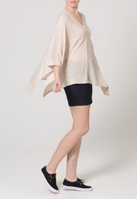 Cardigan lavorato a maglia beige con scollo a V, vestibilità ampia e maniche larghe, abbinato a pantaloni corti in denim scuri e scarpe nere con lacci.