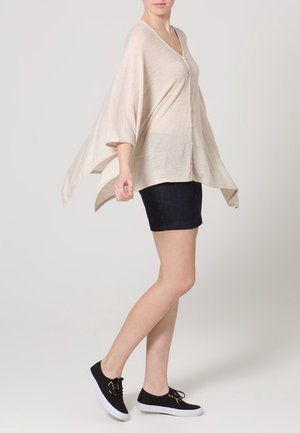 Beige stickad cardigan med V-ringning, lös passform och stora ärmar, parat med mörk denimshorts och svarta snörskor.