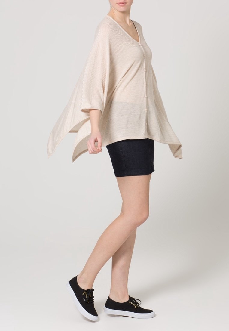 Cardigan lavorato a maglia beige con scollo a V, vestibilità ampia e maniche larghe, abbinato a pantaloni corti in denim scuri e scarpe nere con lacci.