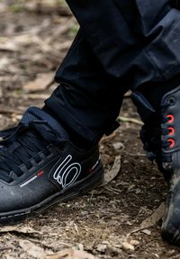 Le scarpe da ginnastica nere presentano un design elegante, una suola in gomma testurizzata, dettagli rossi e un grande logo bianco sul lato, abbinate a pantaloni neri.