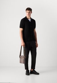 Jonge man in een zwart, gestructureerd overhemd met korte mouwen, zwarte broek, zwarte sneakers, met een kleine taupe schoudertas met hengsel.