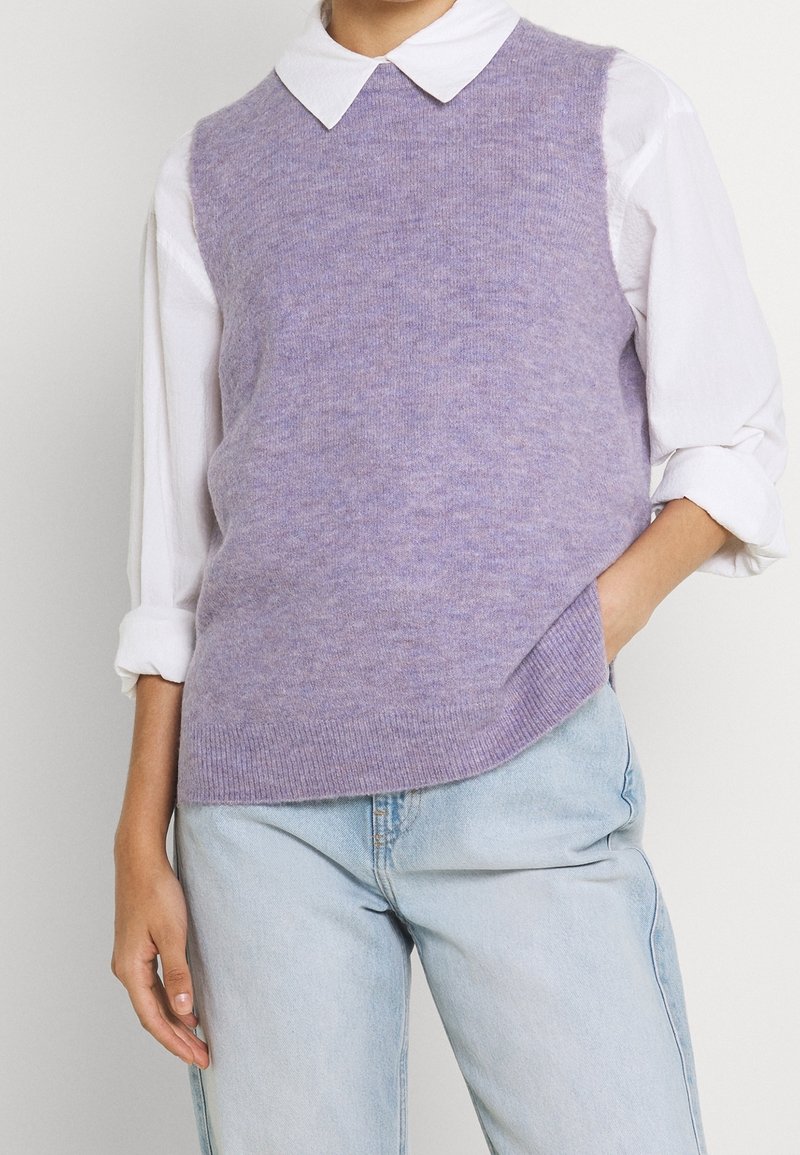 Personne portant un pull sans manches en maille lavande douce sur une chemise blanche à col, avec un jean bleu clair, main dans la poche.