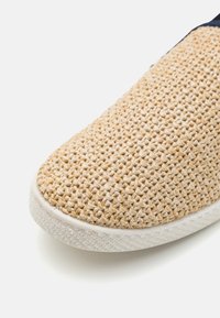 Chaussure à enfiler en paille tissée avec un dessus beige texturé, semelle en caoutchouc blanc et bout arrondi, mettant en avant un aspect en matériau naturel.