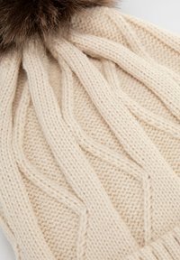 Cappellino lavorato a maglia color crema con motivo a trecce testurizzato. Presenta un morbido pon pon in pelliccia marrone sulla sommità.
