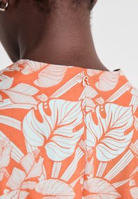 Tissu orange avec des motifs de feuilles blanches, doté d'une fermeture à boutons au dos. Texture lisse et design simple, adapté pour le port décontracté.