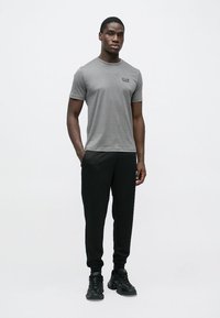 Maglietta grigia con scollo tondo e piccolo logo, abbinata a pantaloni jogger neri e sneakers nere. Design semplice e aderente.