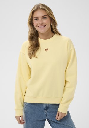 Usmievavá žena s dlhými blond vlasmi, oblečená v svetložltej mikine s výrezom v tvare srdca a modrých džínsach, stojaca pred jednofarebným pozadím.