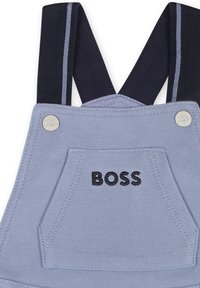 Salopette bleu clair avec des bretelles réglables bleu marine, texte logo noir et une poche avant. Comprend des accents en métal et un tissu texturé.