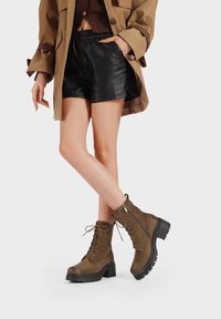 Stivali alla caviglia marroni con lacci, tacco a blocco, materiale in suede texturizzato e dettaglio con zip laterale. Indossati con pantaloni in pelle neri e un cappotto beige.