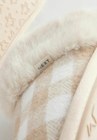 Beige tossut, joissa on pehmeä, karvavuorattu kaulus. Tossuissa on vaaleanruskea ja valkoinen ruudullinen kuvio, ja brändätty logo on näkyvissä. Pintarakenne näkyy pohjassa.