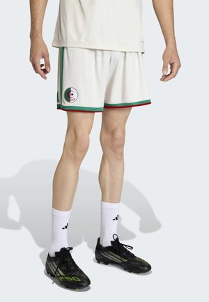 Bílé sportovní šortky se zelenými a červenými akcenty, s kruhovým logem Alžírska. V kombinaci s černými a zelenými kopačkami a bílými ponožkami.