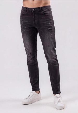 Mand iført slim-fit udfaldne sorte jeans med let slidte detaljer og hvide sneakers står mod en ensfarvet baggrund.