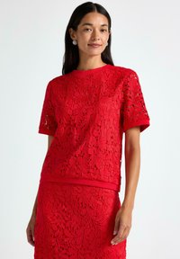 Kvinde med langt, mørkt hår iført sølvøreringe og en matchende rød blonde top med korte ærmer samt nederdel, stående foran en hvid baggrund.