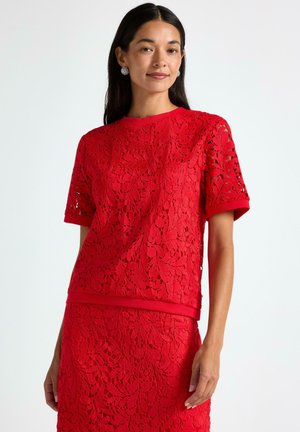 Kvinde med langt, mørkt hår iført sølvøreringe og en matchende rød blonde top med korte ærmer samt nederdel, stående foran en hvid baggrund.