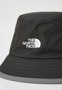 Juodas kibirinis skrybėlė, pagaminta iš lengvos medžiagos, turi pilką akcentą ant krašto ir baltą „The North Face“ logotipą šone.