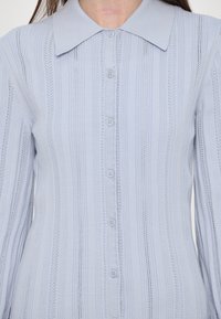Cardigan en maille bleu clair à manches longues avec des rayures verticales ajourées et des boutons devant, porté par une personne aux longs cheveux bruns.