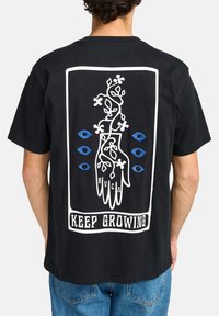 Camiseta de algodón negra con un gráfico blanco de una mano con vides y ojos azules, con "SIGUE CRECIENDO" impreso debajo. Corte relajado.