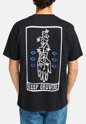 Camiseta de algodón negra con un gráfico blanco de una mano con vides y ojos azules, con "SIGUE CRECIENDO" impreso debajo. Corte relajado.