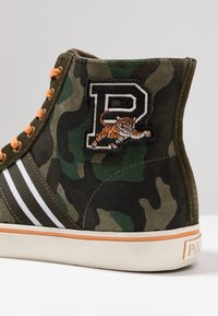 Basket montante avec motif camouflage vert, lacets orange, rayures blanches et tigre brodé au-dessus d'une grande lettre P sur le côté.