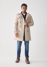 Gabardina beige con botones de doble botonadura, hombreras y bolsillos de hendidura. Llevada sobre un suéter oscuro, combinada con jeans azules y botas marrones.