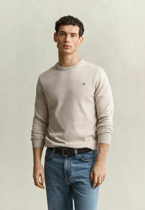 Young man with short dark hair wearing a light beige long-sleeve shirt and blue jeans, standing against a plain light background.