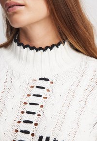 Biały sweter z dzianiny z wysokim, marszczonym czarnym kołnierzem i otwartym wzorem z splotem warkocza; posiada czarne poziome paski i guziki.