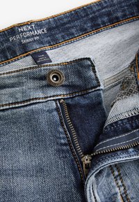 Rifle skinny fit z denimu s čiastočne rozopnutým zipsom, kovovým gombíkom a vnútornou štítkou s nápisom „Next Performance“ a „Made in Bangladesh.“