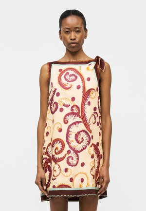 Kobieta w kremowej sukience bez rękawów z dużymi czerwonymi i pomarańczowymi wzorami kwiatowymi i spiralami, zawiązana przy ramieniu, stojąca na jednolitym tle.