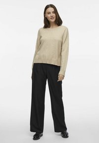 Bež kudumütsiga kampsun ümmarguse kaelusega ja ribakäeservaga, millele on lisatud laia lõikega mustad püksid ja mustad kingad tekstuuriga viimistlusega.