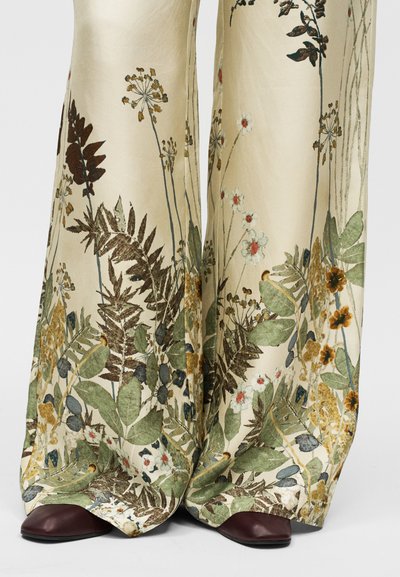 Παντελόνια με φαρδιά γραμμή από ανοιχτό μπεζ ύφασμα με περίτεχνα σχέδια από λουλούδια και φύλλα σε πράσινο, καφέ και κίτρινο, συνδυασμένα με σκούρα μπορντό παπούτσια.