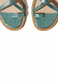 Turkusowe metaliczne sandały z krzyżującymi się paskami, teksturowana powierzchnia, płaska brązowa podeszwa i dekoracyjne metalowe ćwieki wzdłuż krawędzi.