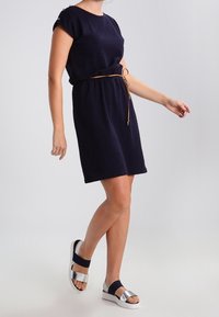 Vestido corto azul marino con tejido texturizado, cuello redondo y mangas enrolladas, ceñido a la cintura con un cinturón marrón claro. Combinado con sandalias plateadas.