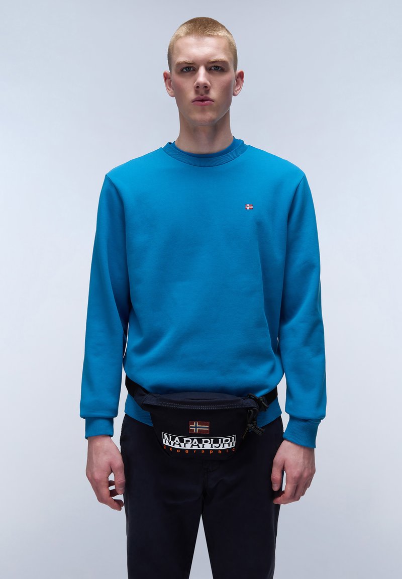 Blaues Sweatshirt mit gerippten Bündchen und Halsausschnitt, kombiniert mit einer schwarzen Gürteltasche mit Logo- und Flaggen-Design, getragen von einem Model.
