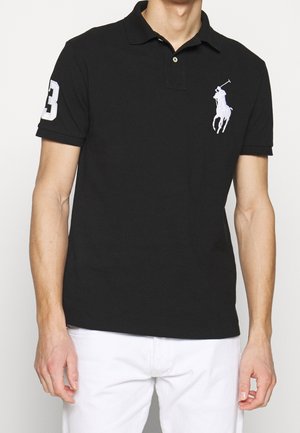Mand iført sort kortærmet poloshirt med hvid polospiller-logo på brystet og nummer 3 på ærmet, kombineret med hvide bukser.