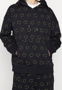 Black hoodie featuring a repeated yellow smiley face pattern, drawstring hood, and a front pocket. Soft fabric and relaxed fit.